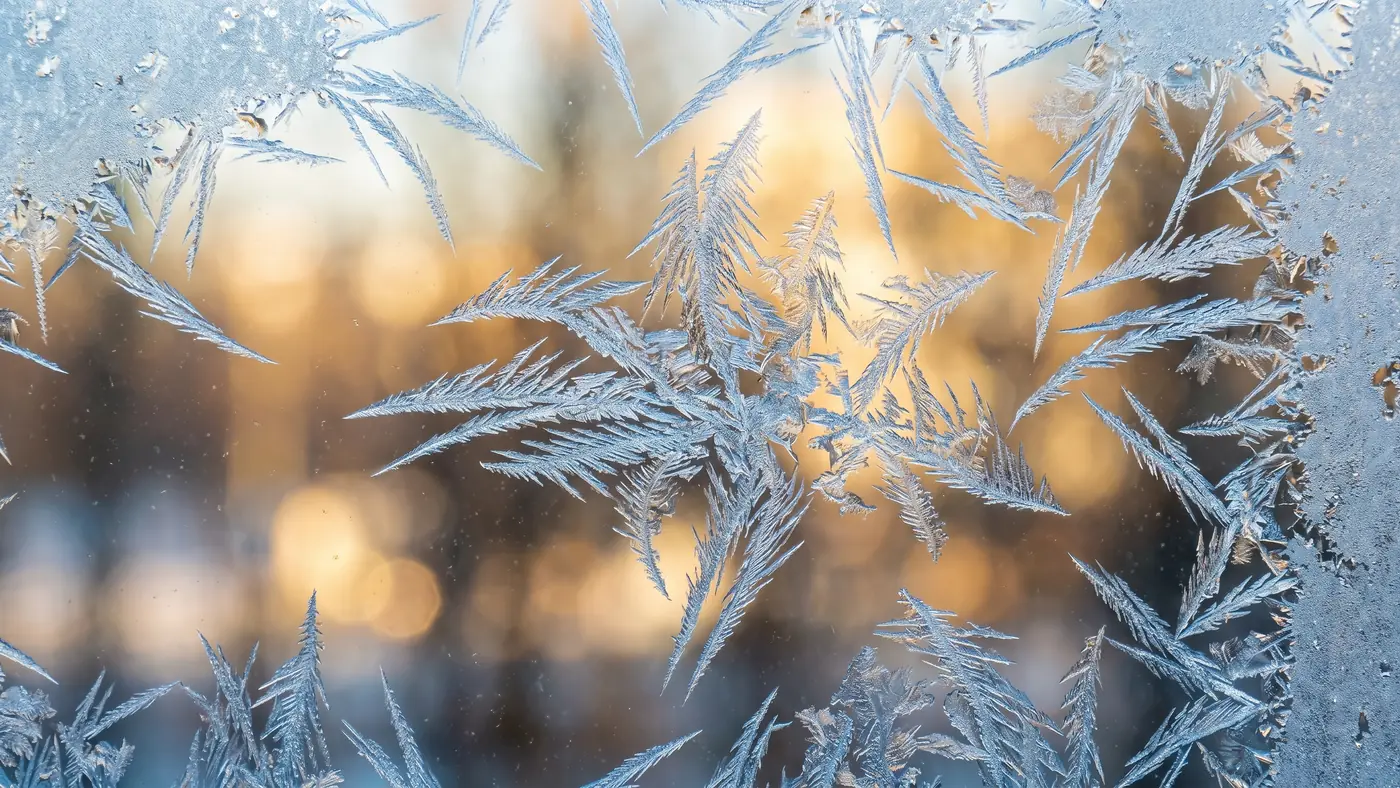 Ice crystals spreading across a windowpane with warm light behind the glass
